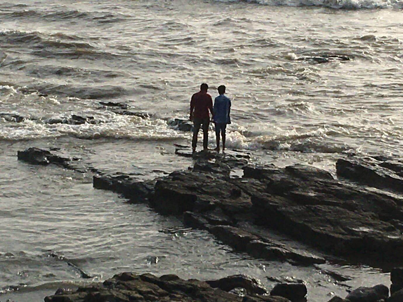 Two men stand hand-in-hand, at the edge of the rocks near Bandra Fort.